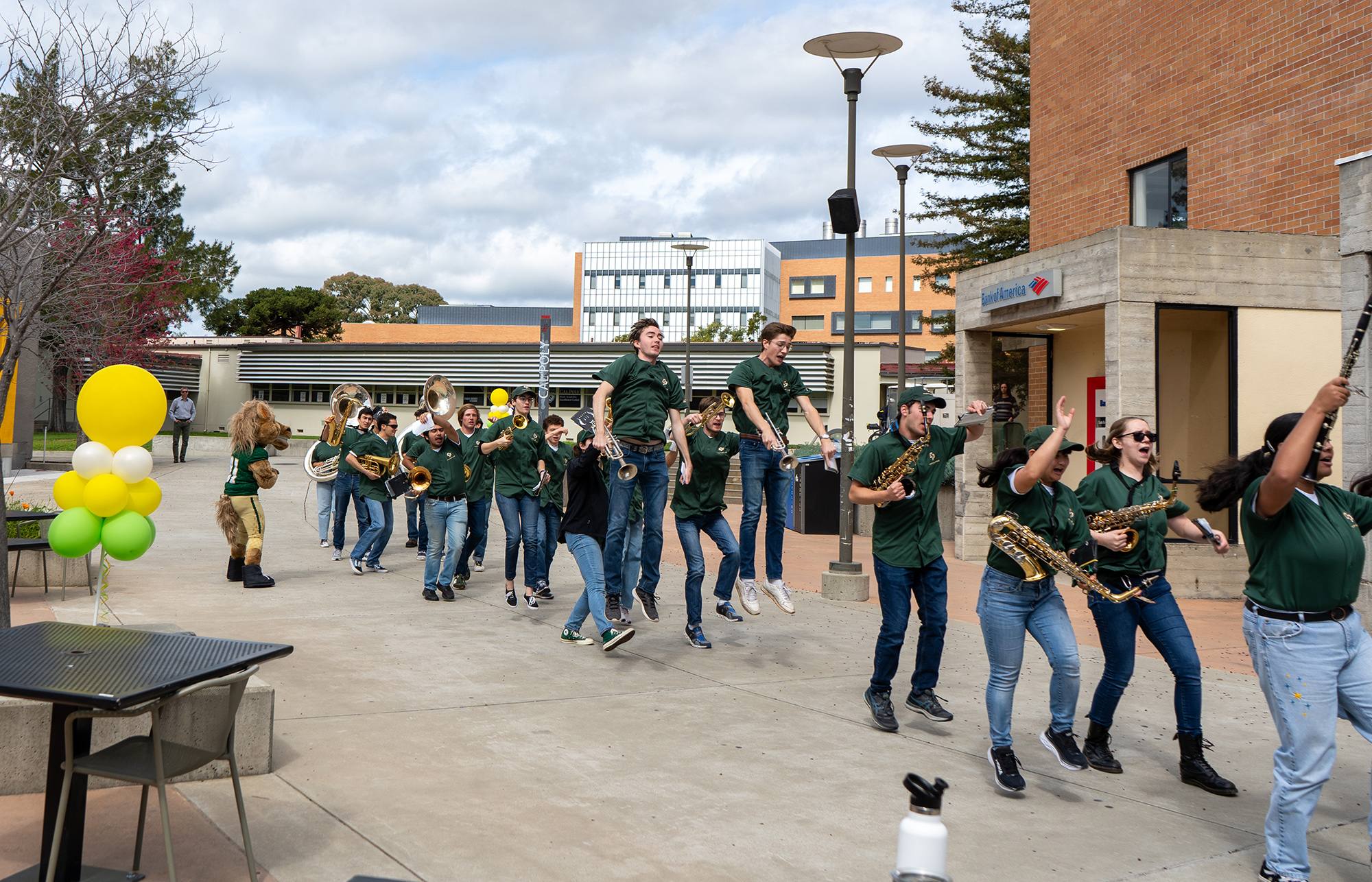 College marching band in green uniforms parades through campus with instruments, accompanied by a mascot and festive balloons.
