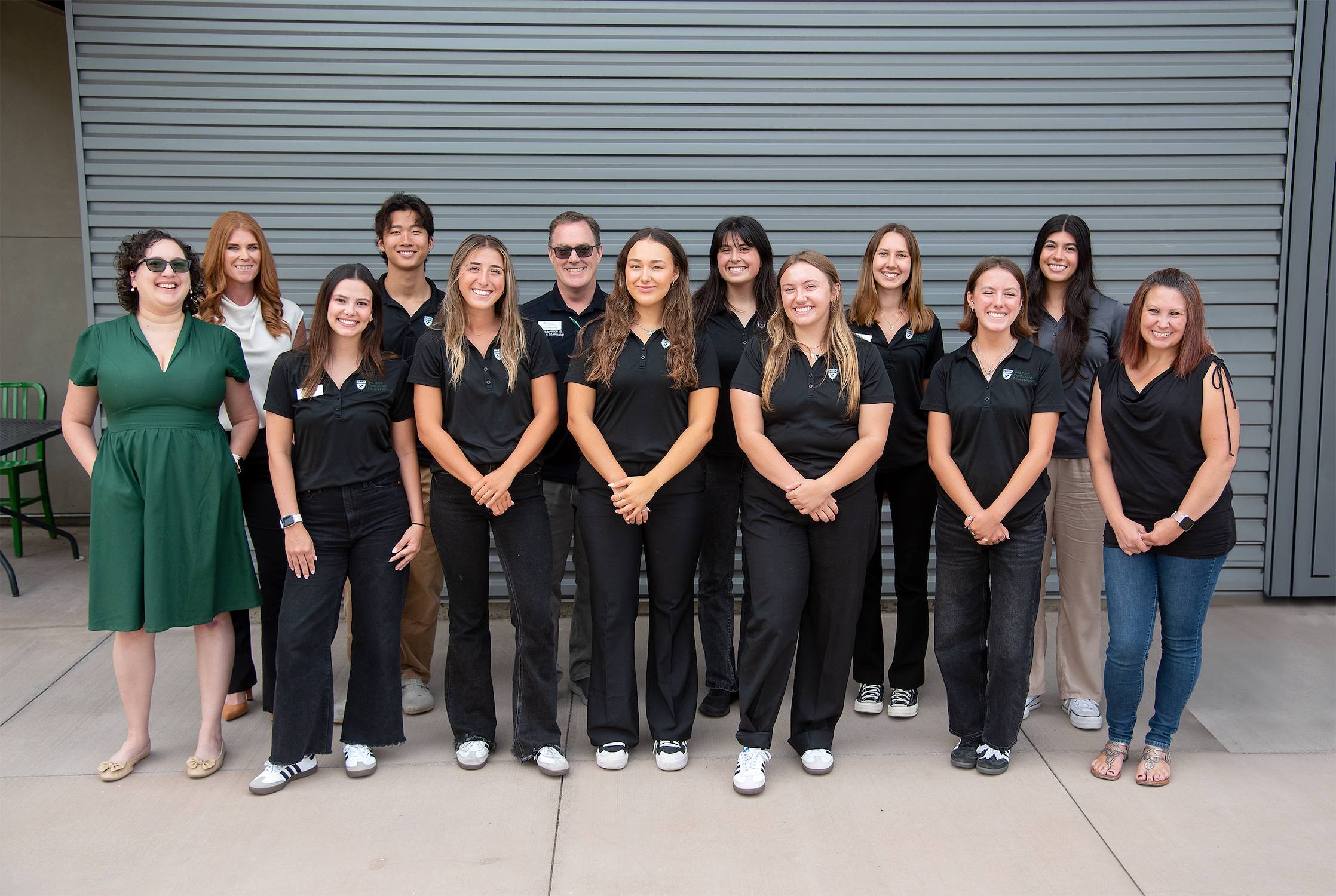 Group photo of a diverse team of students and staff standing in two rows in front of a gray metal wall, with most wearing black Cal Poly attire and smiling at the camera.
