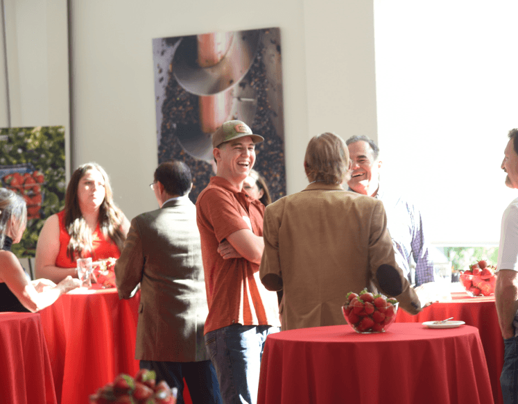 Guests laughing and socializing around red tablecloth cocktail tables with bowls of strawberries at a networking event.