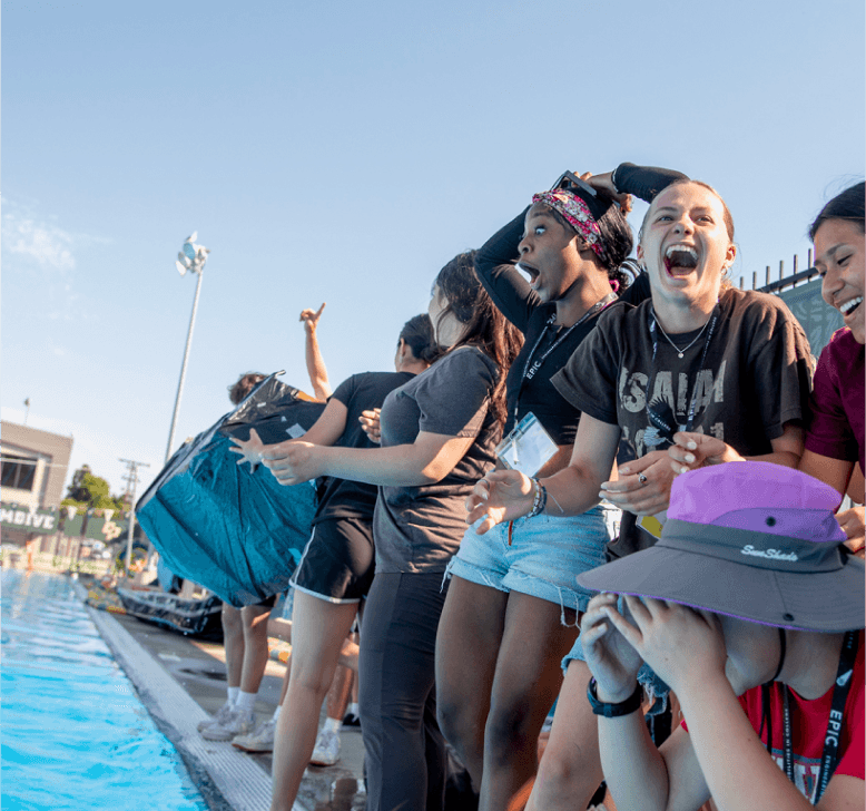 A group of students laughing and cheering poolside during a lively campus event under clear skies.