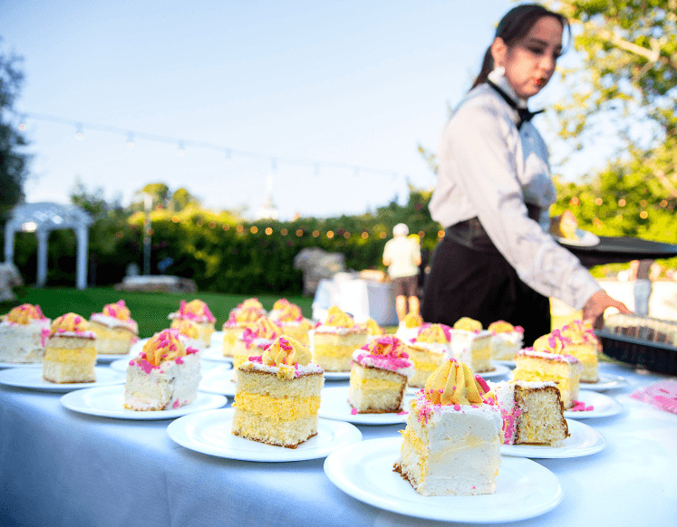 Plated slices of wedding cake with pink and yellow frosting served at an outdoor reception, with a server arranging desserts in the background.