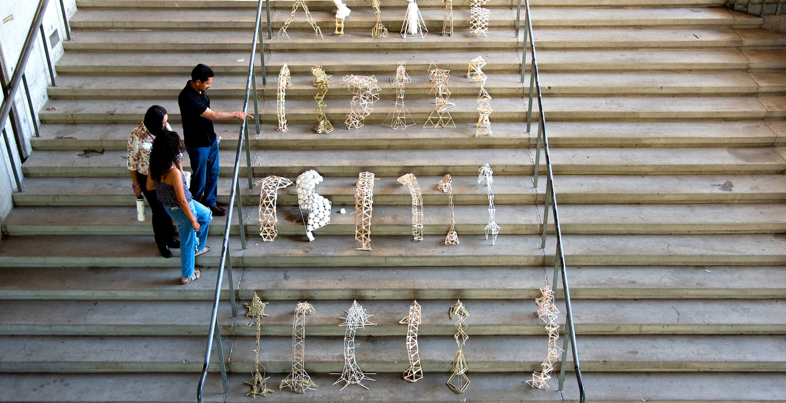 Students evaluating architectural structures made from sticks and string arranged on concrete stairs during a design project showcase.