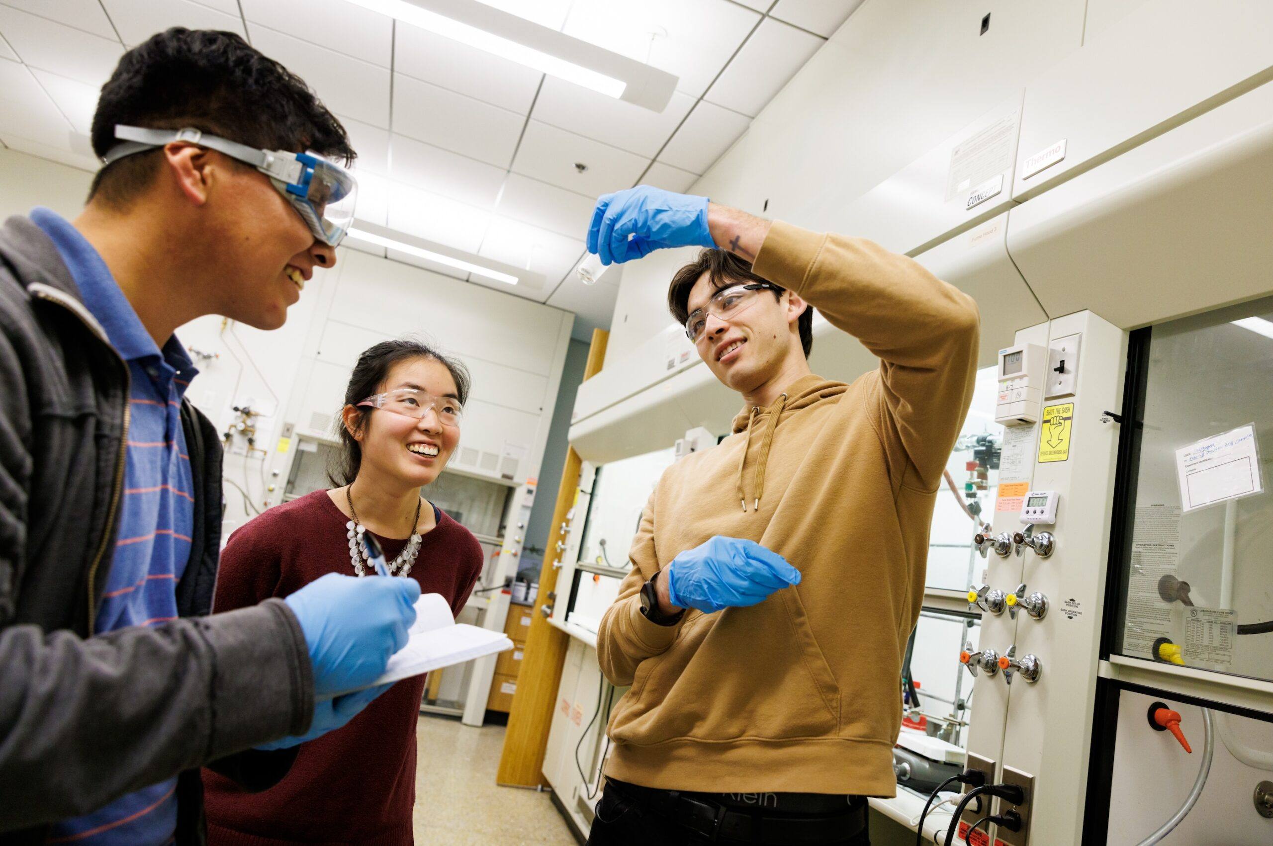 Three college students in safety goggles and gloves conducting an experiment in a university chemistry lab, one holding up a vial while others observe and take notes.