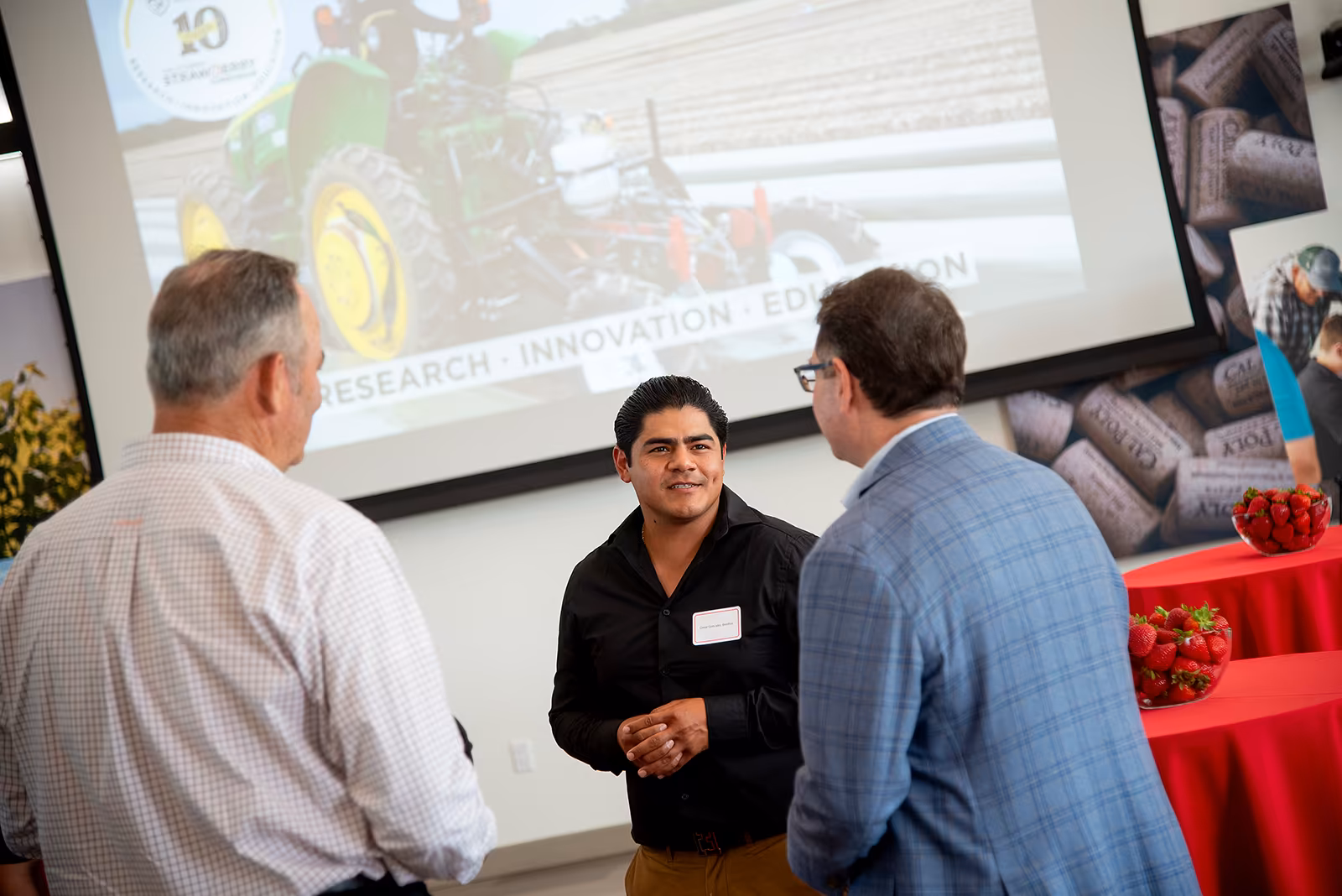 Man networking with two attendees at an agricultural innovation event, with a presentation slide showing a strawberry research project in the background.